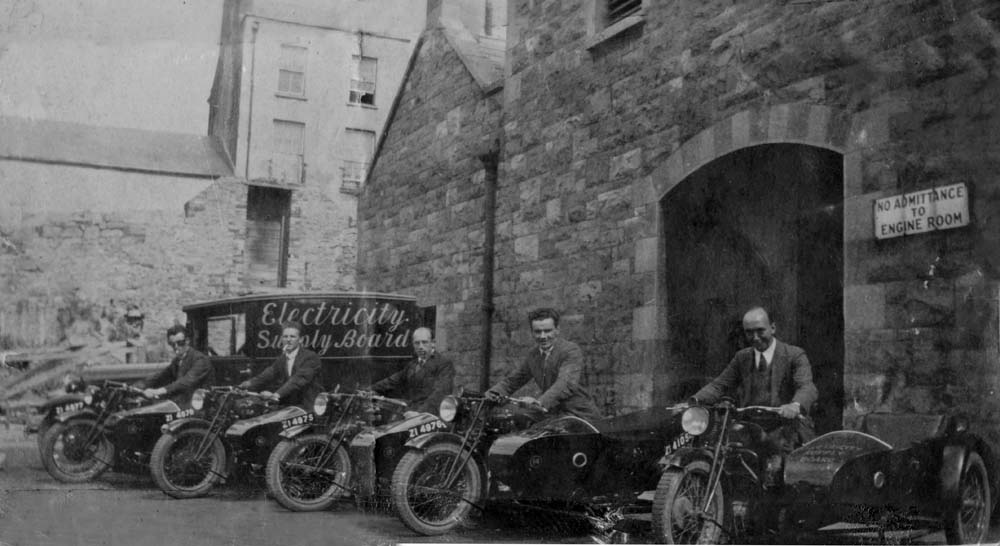 Black-and-white photograph showing a row of Electricity Supply Board motorcycles with sidecars parked outside an industrial stone building.